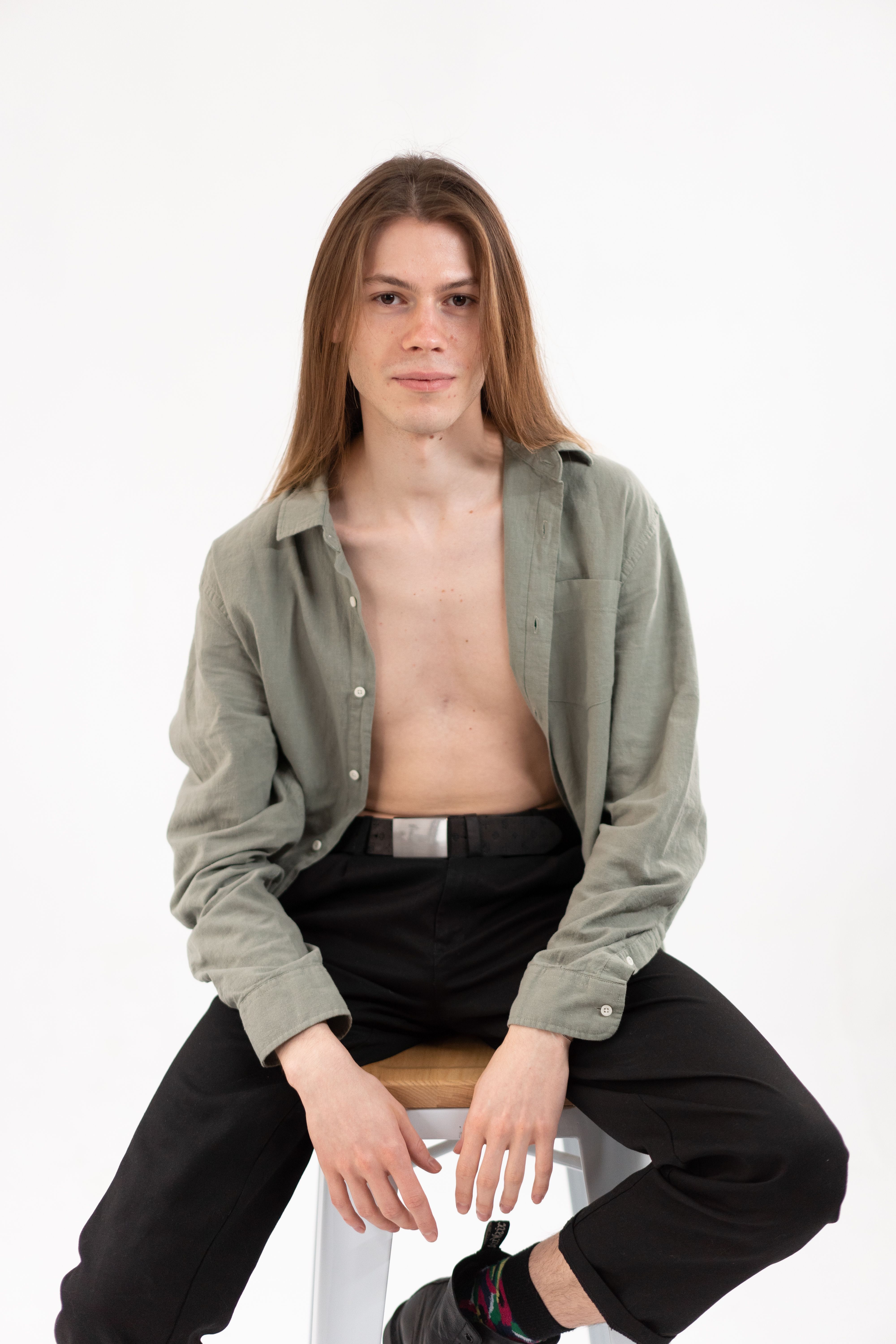 Casual portrait of a young man with long hair seated on a bar stool in a studio setting.