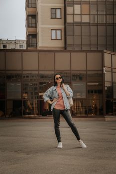 Confident woman in denim jacket and sunglasses posing outdoors against modern building.