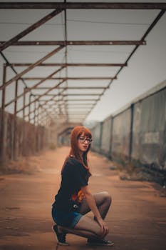 Fashionable woman squatting in an industrial outdoor setting with a metal frame structure.