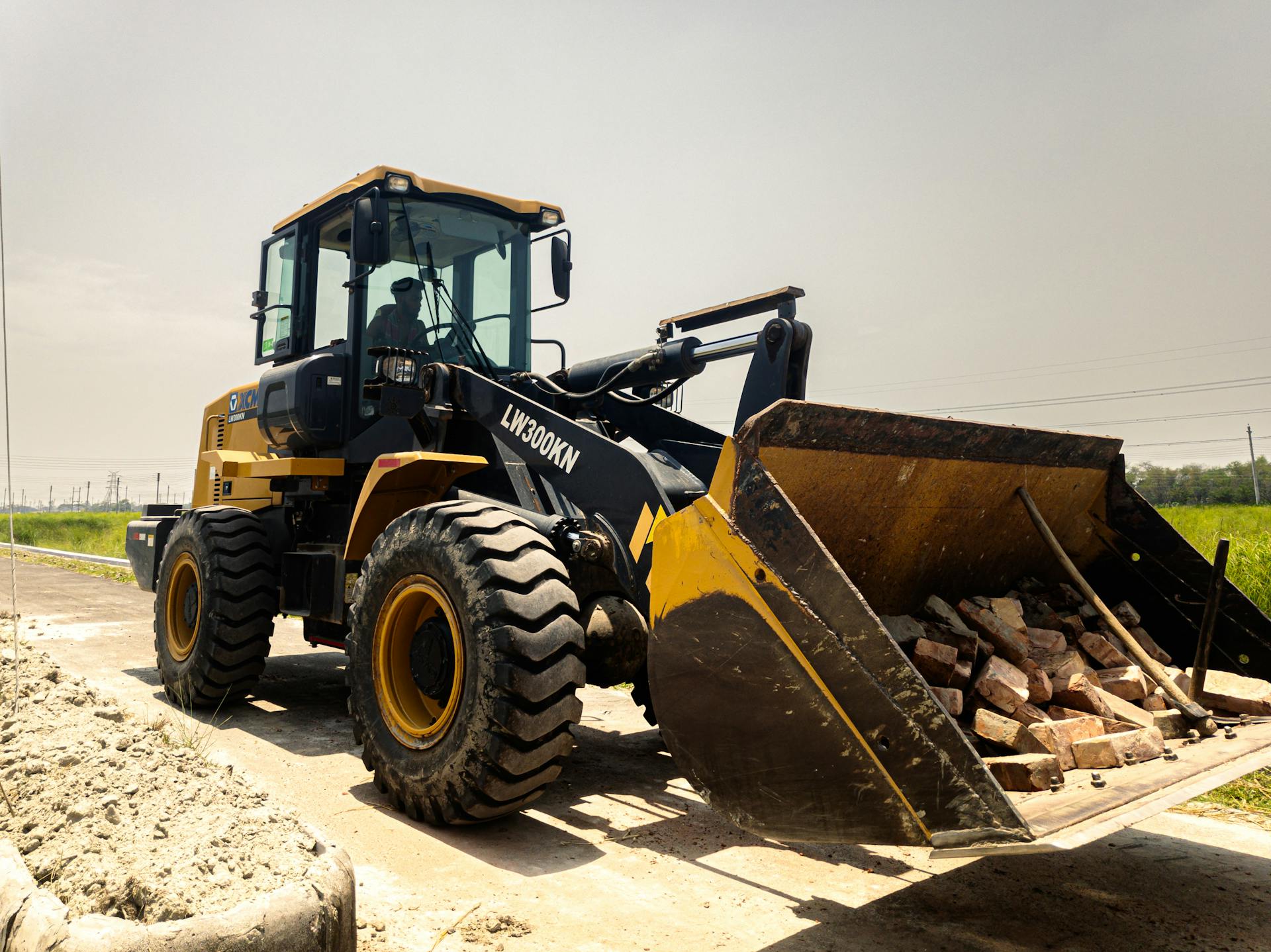 Tractor mounted loader mechanism with heap of bricks in metal bucket on road in countryside near grassy field on summer day