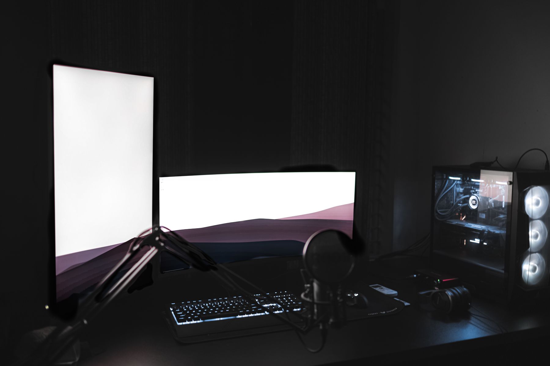 Dark gaming setup with dual monitors, gaming keyboard, microphone and illuminated gaming computer