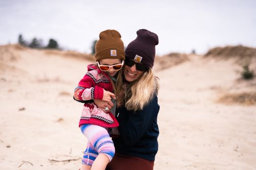 Free Smiling mother and daughter at the beach, bundled in cozy attire and sunglasses, enjoying a playful hug. Stock Photo