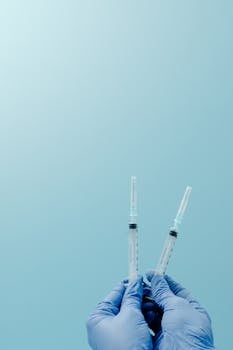 Close-up of gloved hands holding syringes on a light blue background.