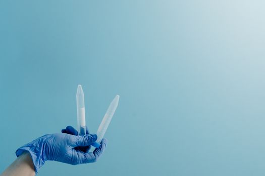 A healthcare worker holds two lab test tubes while wearing blue surgical gloves against a blue background.