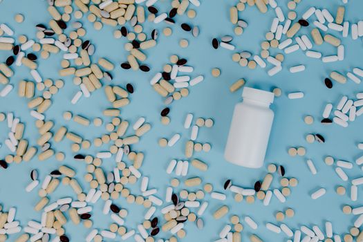 Top view of various pills scattered around a white bottle on a blue surface.