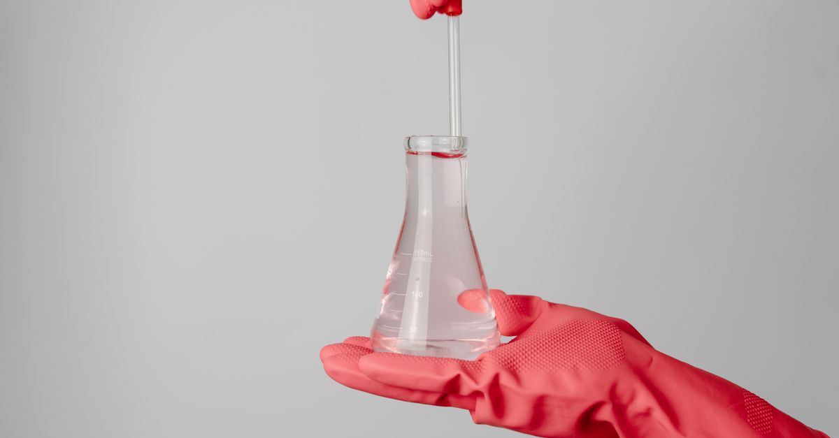 Laboratory scene showing red-gloved hands holding a flask with clear liquid.