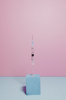 A vertical syringe on a pastel pink background with blue block, perfect for medical themes.