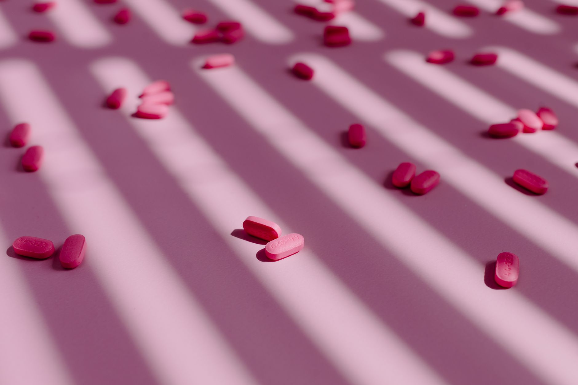 Pink pills scattered on a surface with striped shadows. Minimalist and artistic composition.