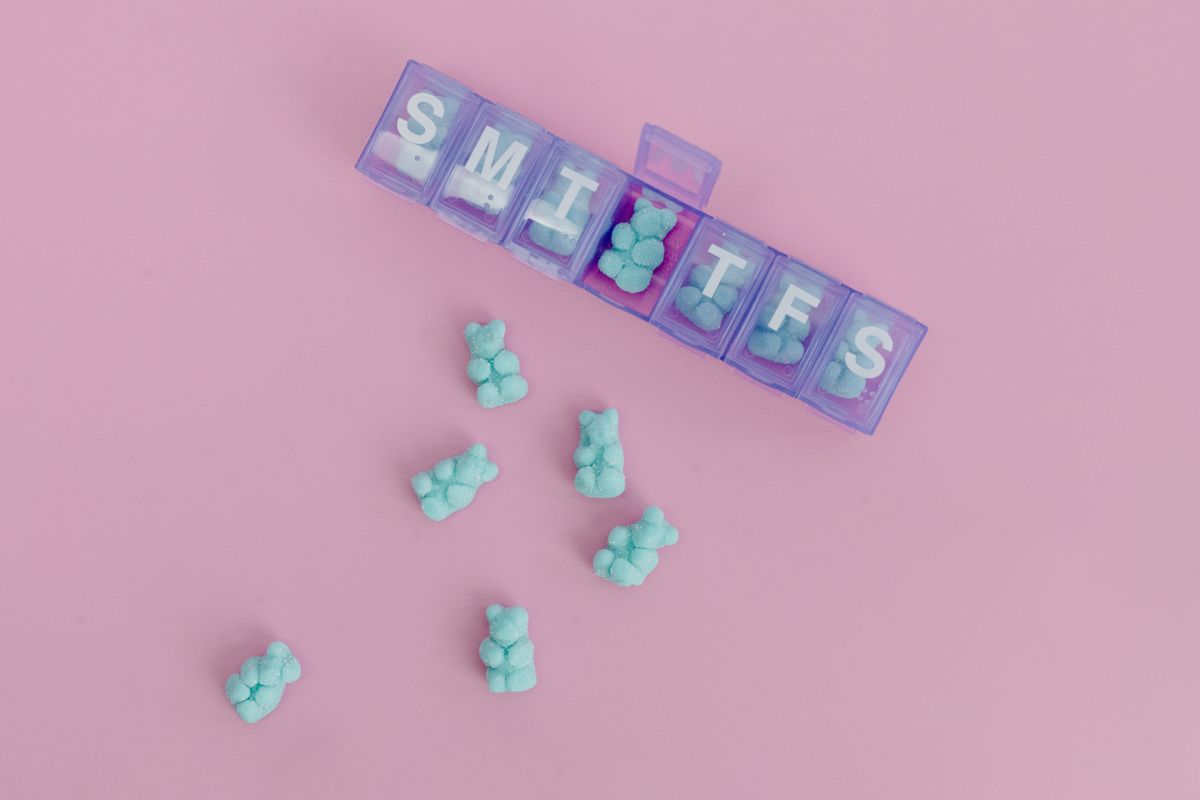 Colorful gummy bears sorted in a weekly pill organizer on a pink background.