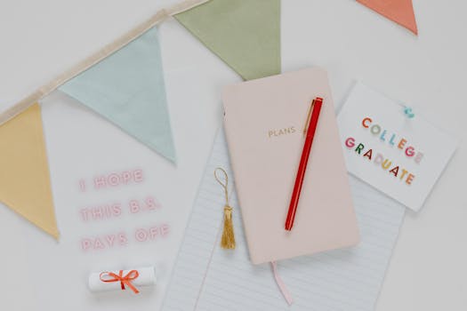 Top view of graduation essentials including a pink notebook, red pen, and celebratory decorations.