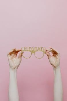 Hands holding glittery celebration party eyeglasses against a pink background.