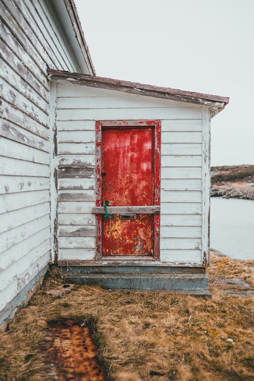 Old white building with shabby red door located on coast with dried grass near lake in countryside against cloudless sky