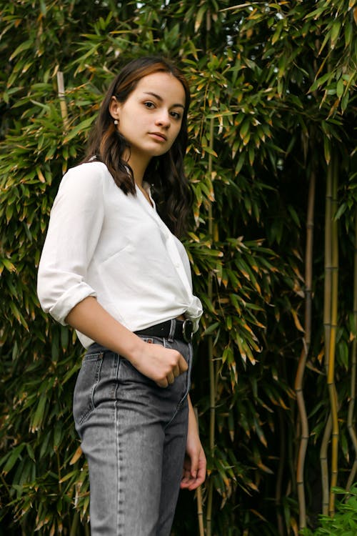 Woman in White Long Sleeve Shirt and Blue Denim Jeans Standing Near Green Plants