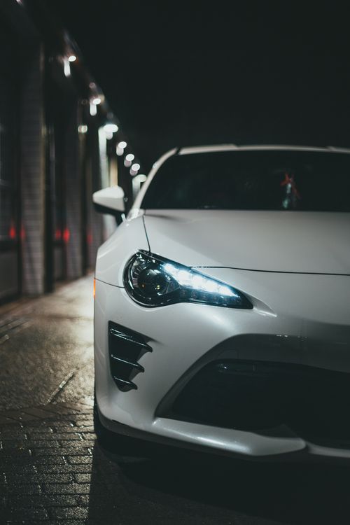 Free Close-up of a white car with illuminated headlight parked on a dimly lit street at night. Stock Photo