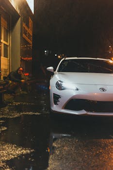 A sleek white car parked on a wet sidewalk at night, reflecting city lights.