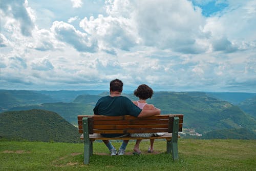 Free Couple Sitting on Brown Wooden Bench Near Mountains Covered With Grasses Under Blue Cloudy Sky Stock Photo
