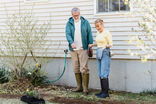 Full length of glad aged couple in rubber boots standing near house wall and looking at greenery on ground