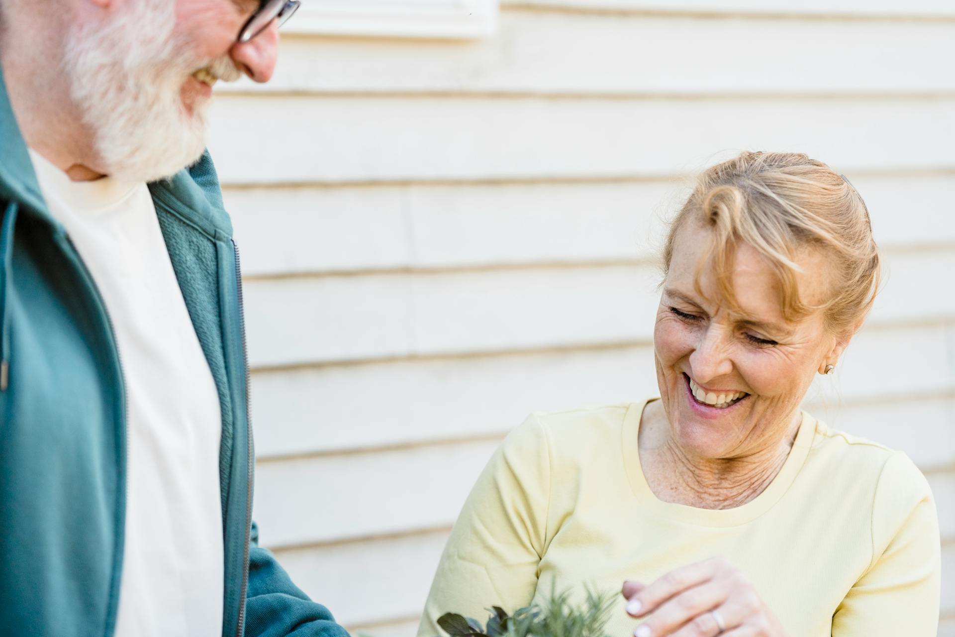 Elderly woman laughing while touching green plants in hands of husband near wall