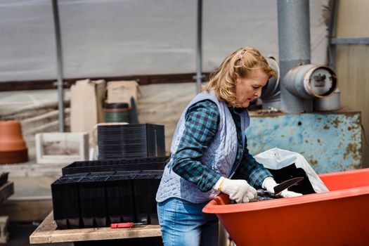 Side view of mature female gardener with shovel standing near cart with soil while working in garden with various boxes