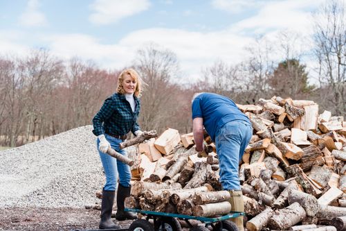 Free An elderly couple working together to stack chopped wood into a wheelbarrow outdoors. Stock Photo