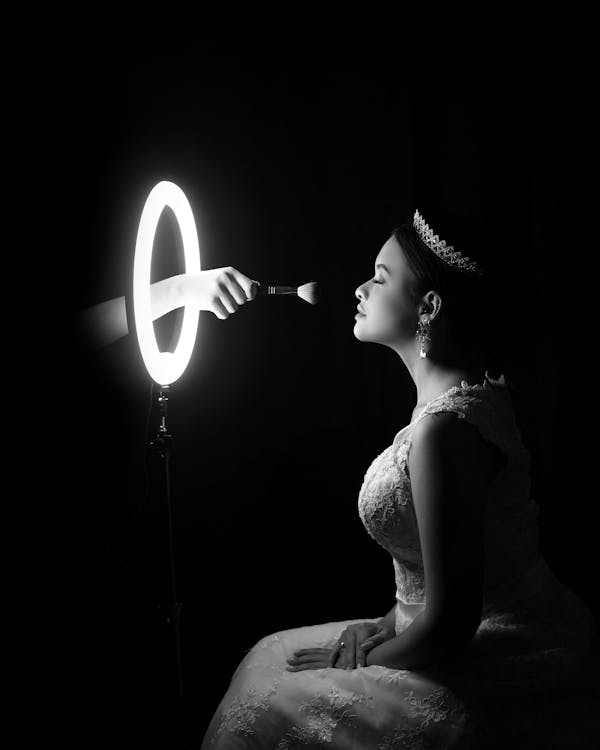 Side view of black and white unrecognizable beautician applying makeup on face of gorgeous bride in elegant dress and tiara sitting in dark room in front of round illuminated mirror