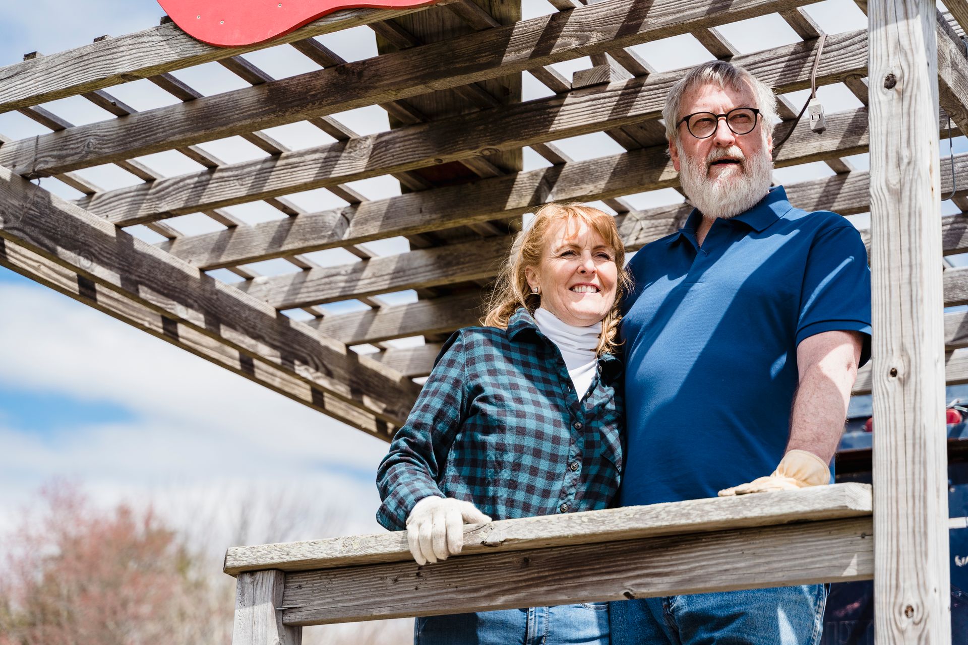 Smiling senior couple stands together outdoors under a wooden pergola, embracing life and love.