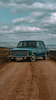 A classic vintage car parked on a scenic dirt road in Uşak, Türkiye under a cloudy sky.