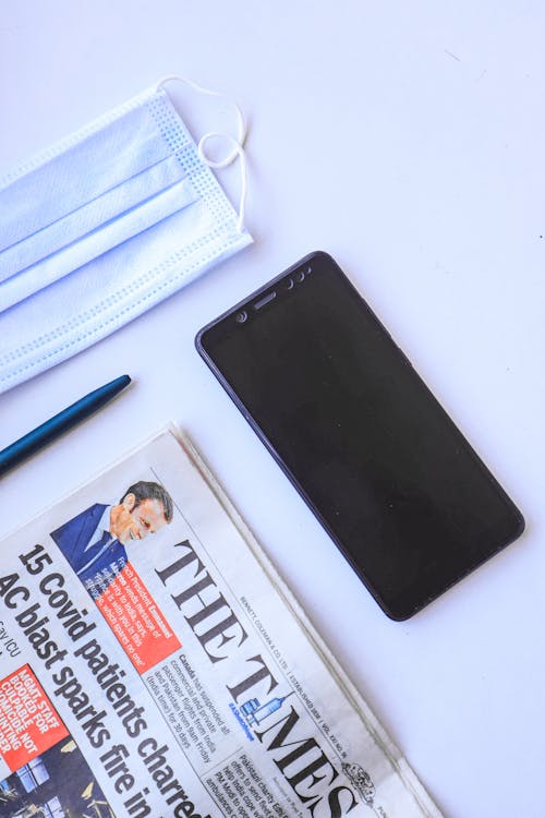 A Smartphone and Newspaper Near a Pen and a Facemask