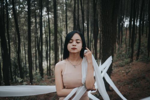 Free A serene woman stands in a forest with eyes closed, wrapped in white cloth, exuding calmness and connection with nature. Stock Photo