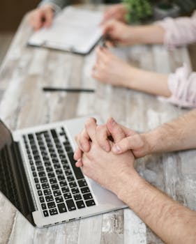 Close-up of hands during a business meeting with a laptop and documents on a wooden table.