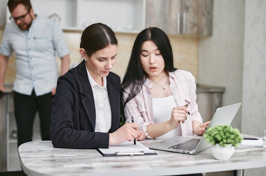 Two businesswomen collaborating on a project with a laptop and documents, working in a modern office environment.