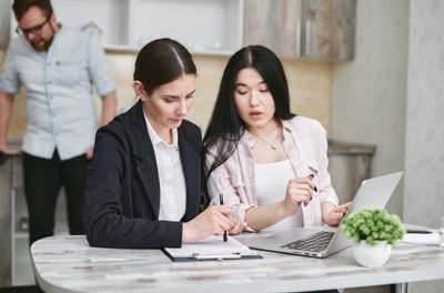Cũng hai người phụ nữ đó tại một chiếc bàn, một người đang viết vào bảng kẹp giấy, một chiếc laptop mở bên cạnh