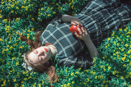 A woman lies in flowers holding a pomegranate, eyes closed, serene scene.