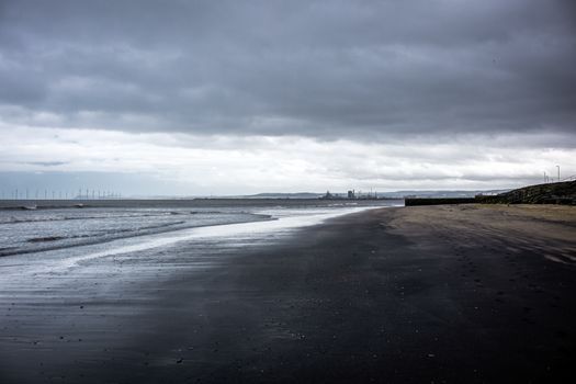 Moody beach scene with dark clouds and distant offshore wind turbines under an overcast sky.