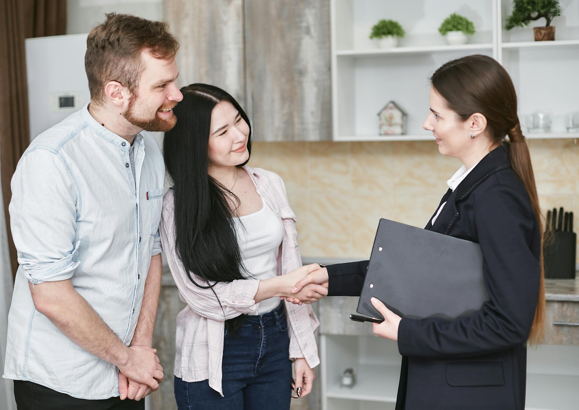 A real estate agent greets a couple with a handshake in a modern home interior.