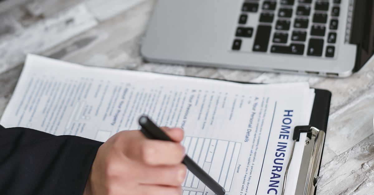 An adult reviews and signs a home insurance policy document on a clipboard next to a laptop.