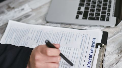 An adult reviews and signs a home insurance policy document on a clipboard next to a laptop.