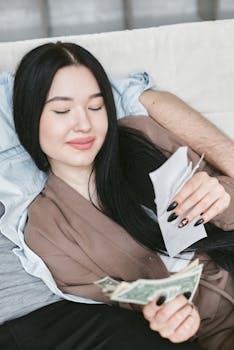 Asian woman lounging indoors, holding money and an envelope, appearing content.