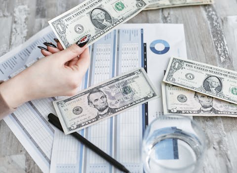 Close-up of a hand holding a dollar bill over financial documents and charts on a wooden table.