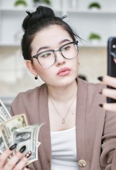 Asian woman with glasses holding dollar bills, taking a selfie indoors.