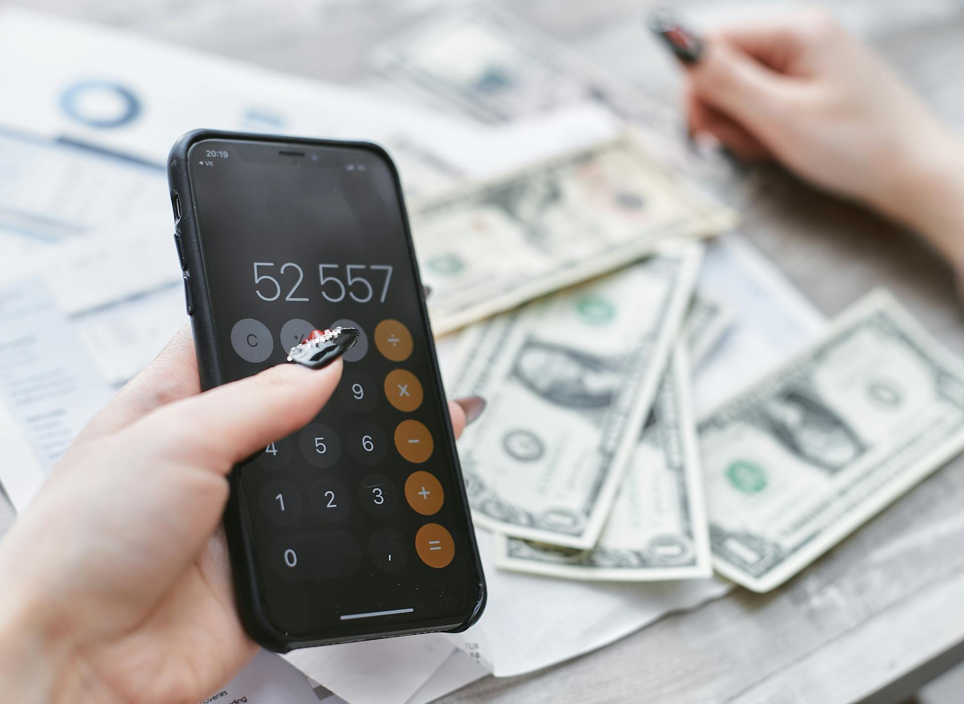 Close-up of hands using smartphone calculator next to dollar bills on table. Financial planning concept.