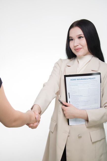 Asian woman in office attire shaking hands, holding home insurance policy document.