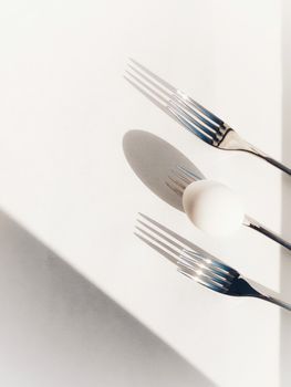 Artistic flatlay of an egg and three forks casting shadows on a white surface, top-down view.