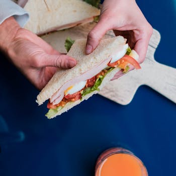 Close-up of a hand holding a fresh sandwich with vegetables and meat on a blue table.