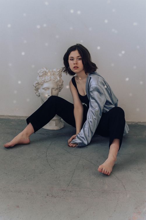 Free Woman in elegant satin blazer sitting barefoot beside Greek sculpture indoors. Stock Photo