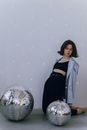 Full body of barefoot female in top and shirt sitting while bending back near silver disco balls on white background with glares