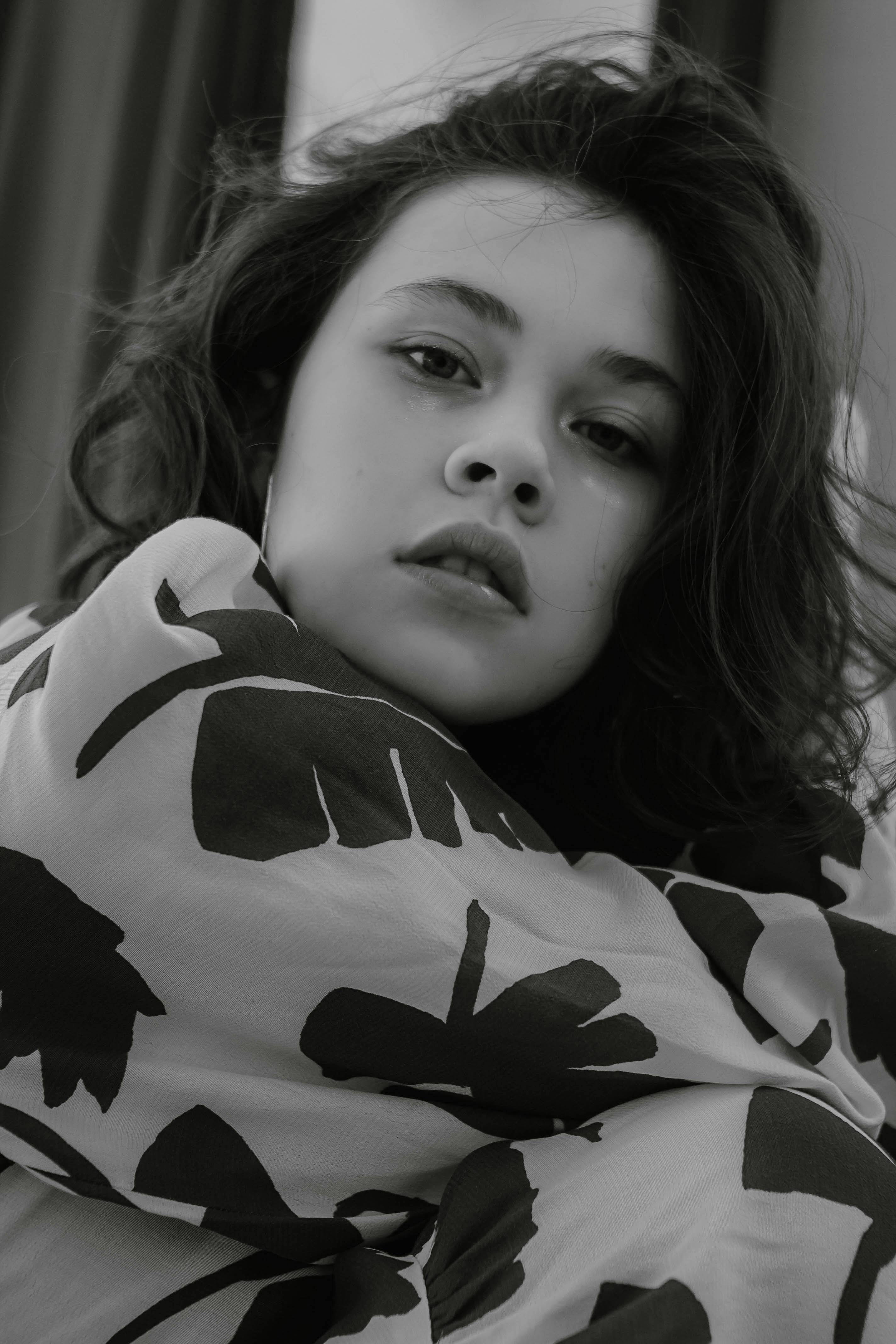 Black and white of calm female with dark hair wearing clothes with ornament looking at camera while sitting in light room at home