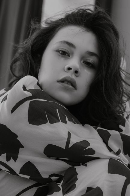 Black and white of calm female with dark hair wearing clothes with ornament looking at camera while sitting in light room at home