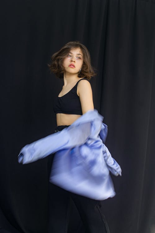 Free Serious female with dark hair wearing top looking away while taking off blurred shirt while standing on black background in modern studio Stock Photo