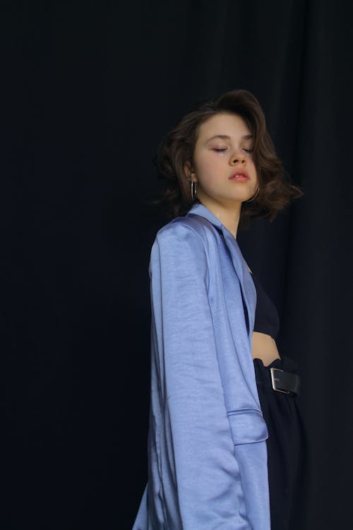 Peaceful young female with dark hair wearing blue shirt while standing on black background with closed eyes in modern studio